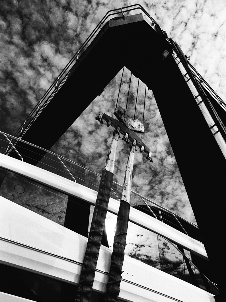 Black and white photo looking up at a large yacht suspended by straps from a boat hoist against a cloudy sky.