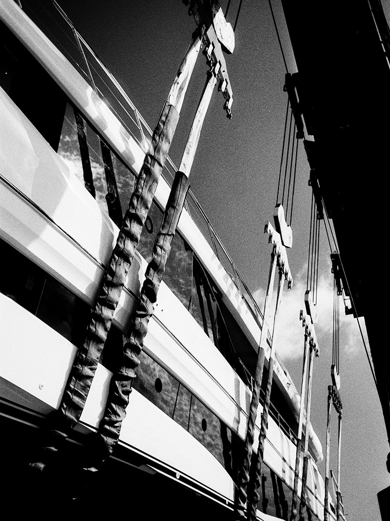 Black and white photo of a yacht being lifted by wide straps on a boat hoist, viewed from below the deck line.