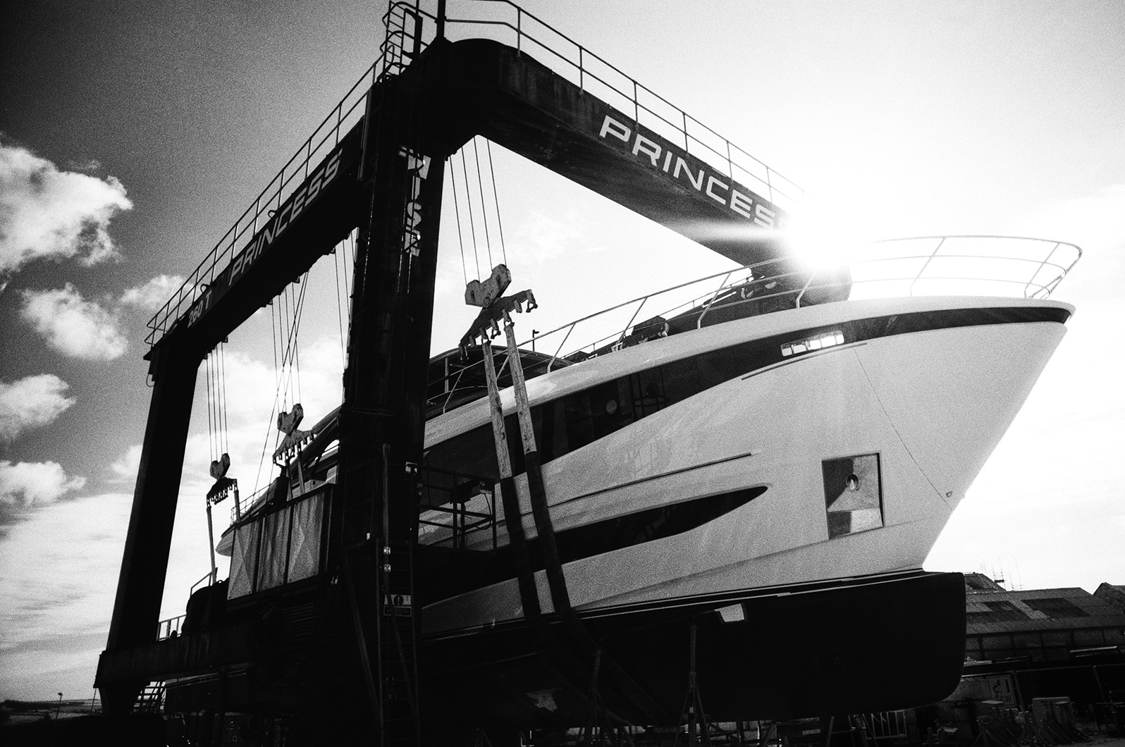 Black and white photo of a yacht in a large boat hoist marked 'Princess', with the sun shining behind the bow.