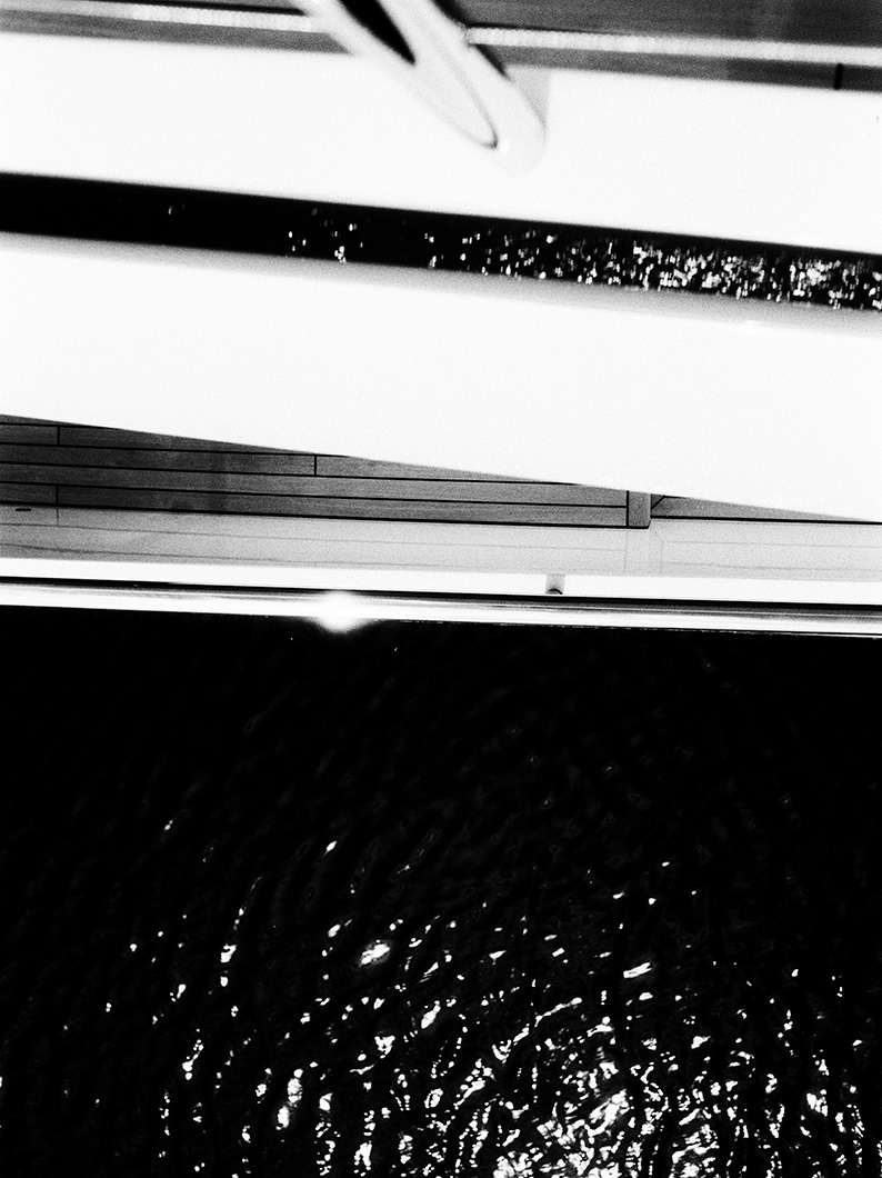 Black and white downward view from a yacht over the deck and railings to the dark rippling water below.