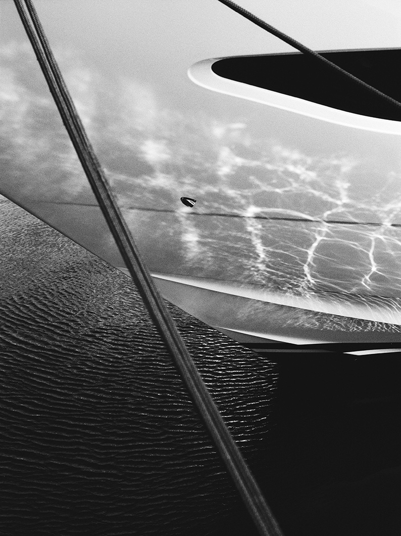 Black and white photo of a yacht bow above rippling water, with light reflections and mooring lines visible.
