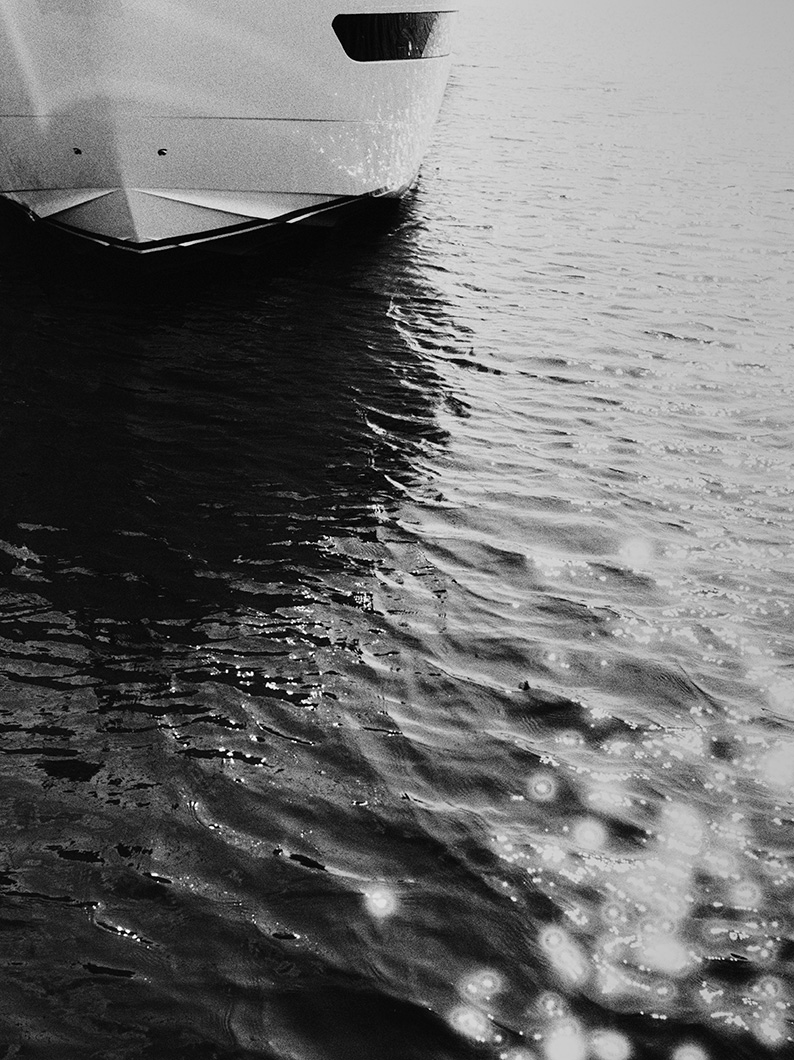 Black and white photo of a yacht bow on the water, with sparkling sunlight reflecting across the ripples.