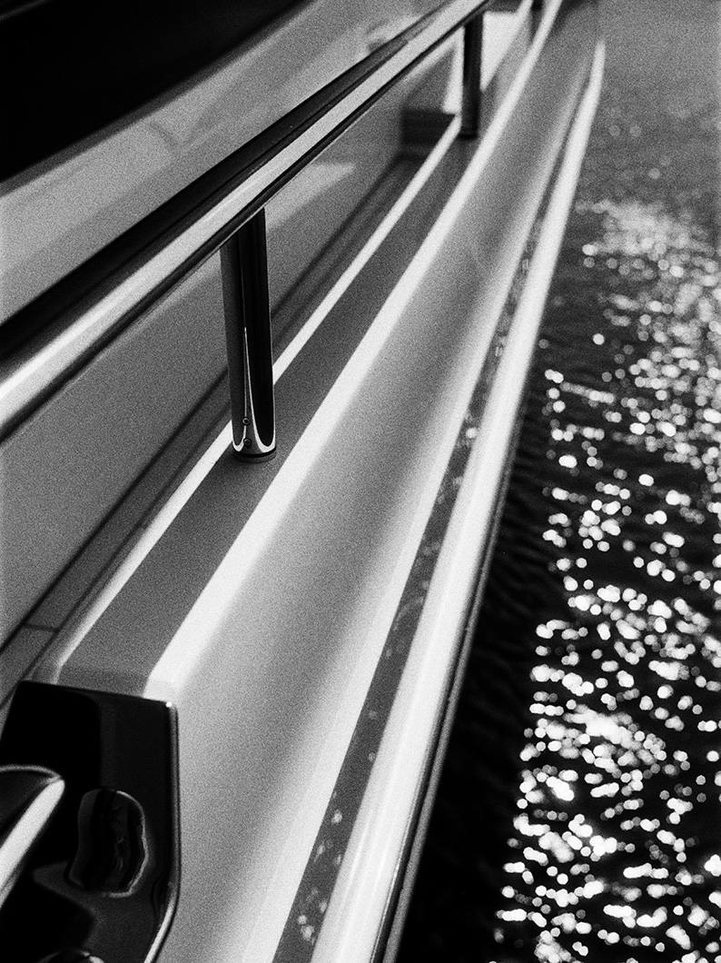 Close-up black and white image of a yacht’s railing and side, with sparkling reflections on the water below.