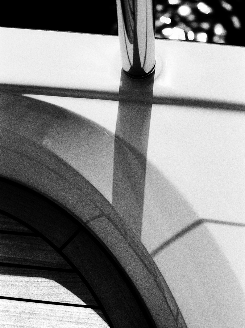 Black and white close-up of a stainless-steel rail stanchion casting a long shadow across white moulding and a curved teak step.