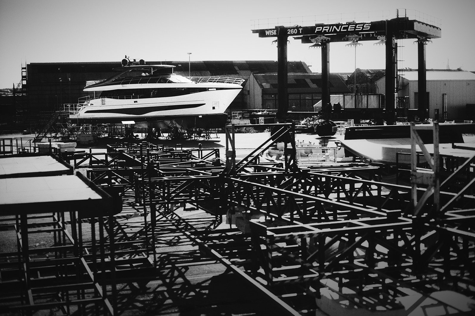 Black and white photo of a yacht on land beside a large Princess boat hoist, with metal scaffolding structures in the foreground.