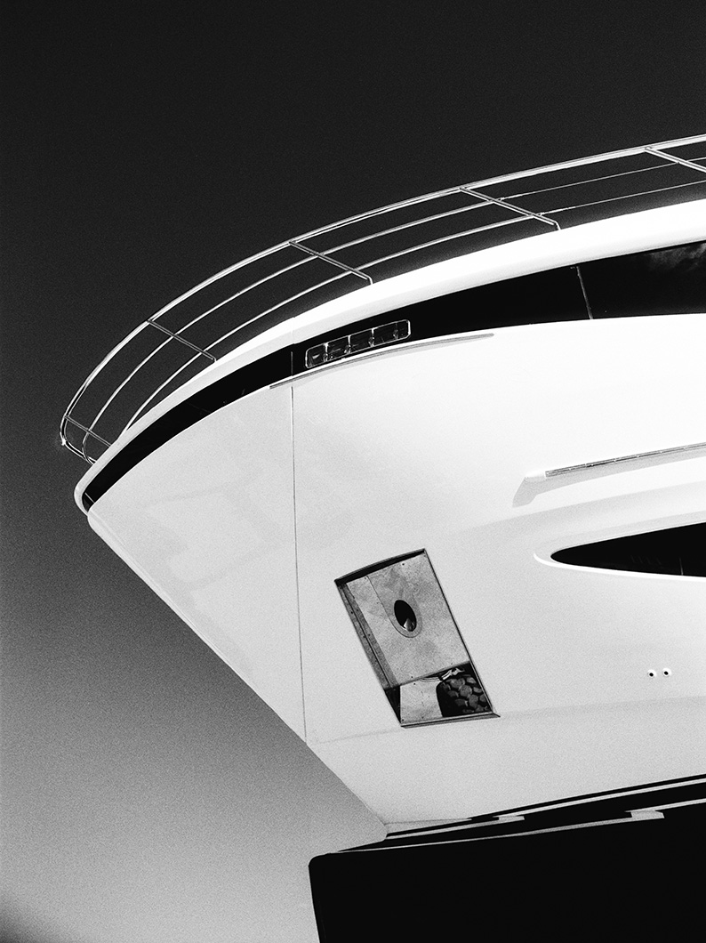 Black and white close-up photo of the bow of a yacht, showing its sleek lines and railings against the sky.