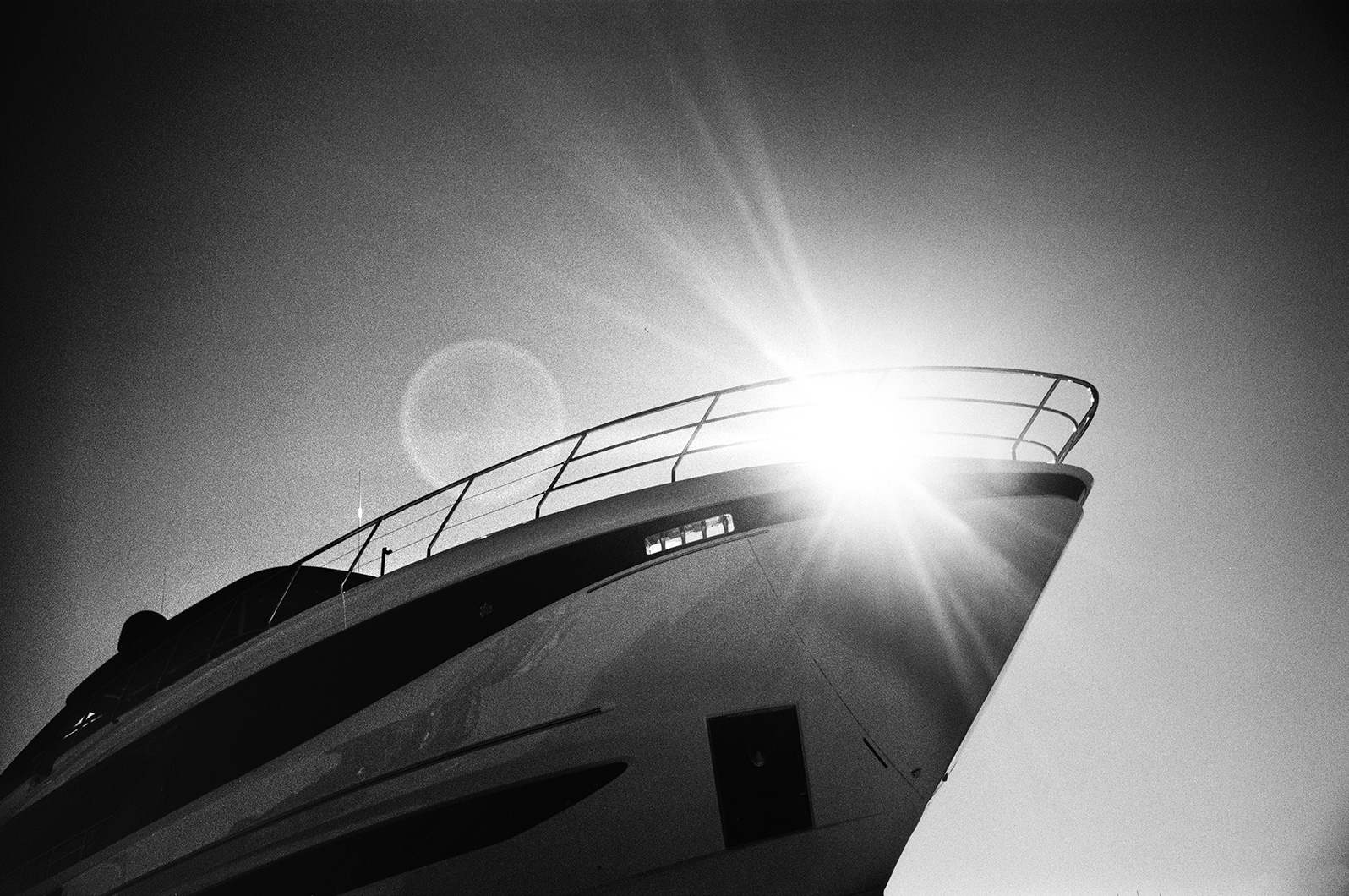 Black and white photo of a yacht’s bow with the sun shining brightly behind it, creating a lens flare in the sky.