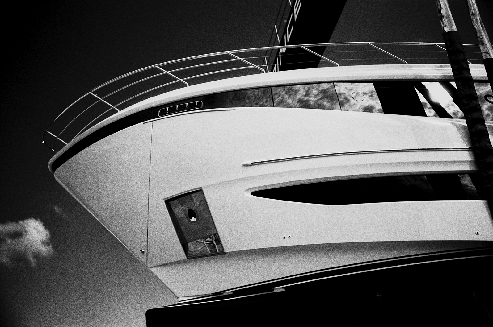 Black and white close-up of a yacht’s bow and railings, with reflections in the windows and straps from a boat hoist visible.
