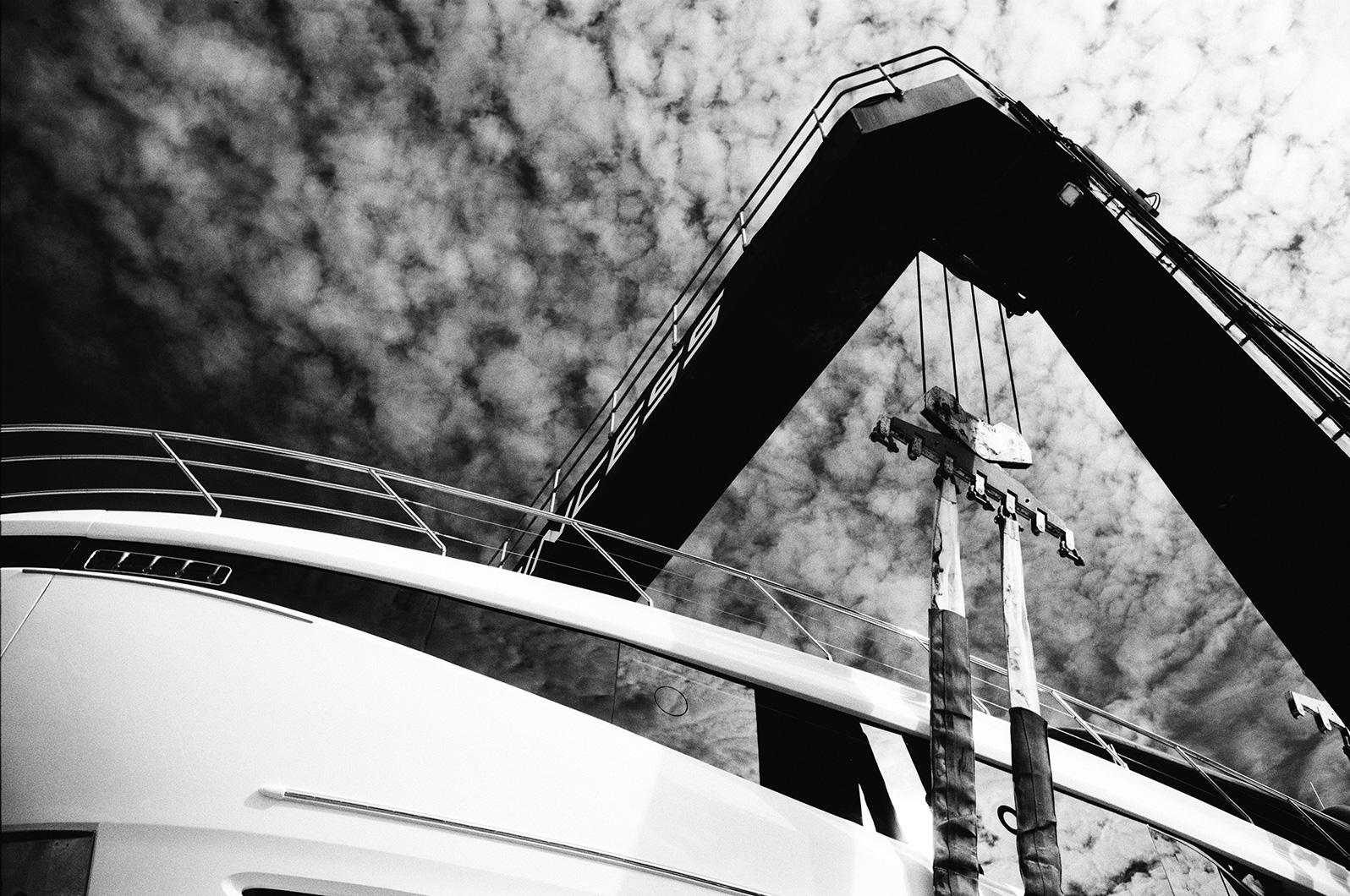 Black and white photo of a yacht in a boat hoist, viewed from below with straps hanging against a dramatic cloudy sky.