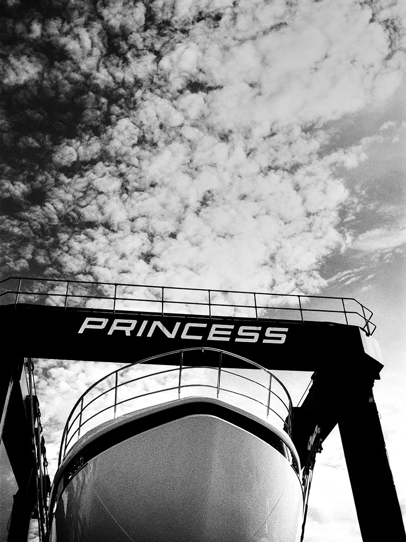 Black and white photo of a yacht’s bow directly beneath a Princess boat hoist, set against a textured cloudy sky.