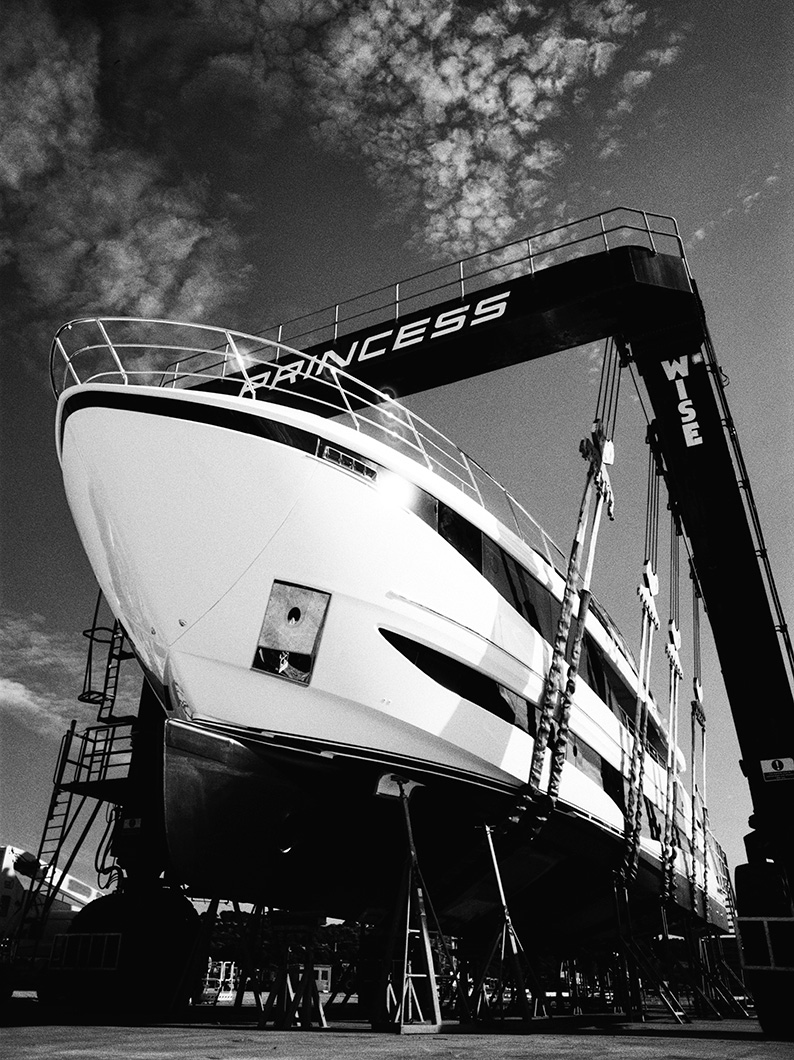 Black and white photo of a large yacht suspended in a Princess boat hoist, with its bow and hull supported by straps and stands.