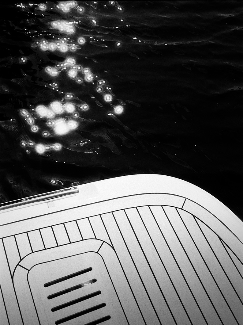 Black and white close-up of a yacht swim platform with sunlight sparkling on the dark water beside it.