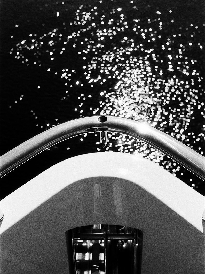 Black and white photo from the bow of a Princess yacht, with stainless steel railings in the foreground and sunlight glittering on the water.